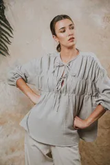 Camisa de algodón con textura arrugada, cuello redondo con cierre de cordones y mangas largas con puños elásticos. Presenta un diseño holgado con frunces en el pecho.