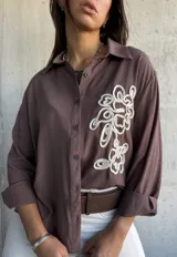 Camisa de manga larga color chocolate, con cuello clásico y cierre frontal de botones. Presenta un bordado decorativo en color crudo en el frente con diseño de formas orgánicas.