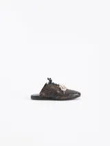 Zapatilla tipo mule (sin talón) de cuero vacuno con efecto gastado en tonos marrones oscuros y negro. Presenta un diseño con recortes y un logo estilizado grabado en el empeine. Tiene cordones blancos y un detalle de pasador metálico en la cordonera.