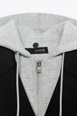 Abrigo de paño negro con diseño cruzado, cierre frontal con cremallera y botones, y capucha gris desmontable con cordones ajustables. Presenta solapas clásicas, manga larga y bolsillos delanteros con vivo.