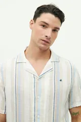 Camisa de manga corta con cuello cubano, confeccionada en una mezcla de lino y algodón con textura. Presenta un diseño a rayas verticales en tonos beige y celeste sobre fondo blanco, con cierre frontal de botones y logo bordado en el pecho.