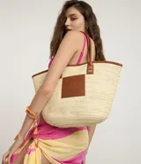 Bolso de playa tipo tote bag, tejido en fibra de papel color natural, con bordes y detalles en cuero sintético marrón. Posee asas de hombro en cuero sintético marrón y un parche cuadrado con logo grabado en el frente.
