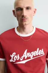Camiseta roja de corte regular fit, con cuello redondo y mangas cortas. Presenta un estampado frontal con la frase "Los Angeles" en blanco y acabados de vivo en contraste blanco en cuello y mangas.