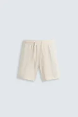 Bermuda de calce regular, color beige claro, confeccionada en tejido de mezcla de algodón con textura jacquard. Presenta cintura elástica ajustable con cordón, bolsillos frontales y un bolsillo trasero de vivo.