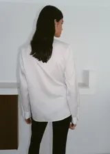 Camisa blanca de manga larga con cuello camisero, cierre frontal de botones y un llamativo detalle de nudo drapeado en el lateral delantero.