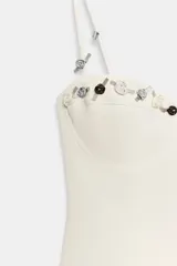 Vestido mini de corte corsetero en color crudo, confeccionado en tejido elástico, con escote corazón, aros en el busto y tirantes finos. Presenta delicadas aplicaciones de pequeñas flores a lo largo del escote y los tirantes.