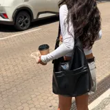Cartera tipo hobo de color negro, confeccionada en material sintético con acabado liso. Presenta un diseño estructurado con dos bolsillos frontales funcionales y un detalle metálico central con el logo de la marca. Cuenta con una correa ajustable con hebilla y cierre principal.