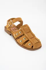 Sandalia tipo cangrejera de gamuza color camel, con diseño de tiras entrelazadas y tachas metálicas doradas. Cuenta con cierre de pulsera con hebilla ajustable y suela plana de goma.