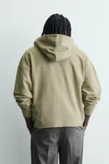 Sudadera de corte relajado color verde oliva con efecto lavado. Presenta capucha con abertura frontal, bolsillo tipo canguro, mangas largas con puños de rib y bajo con acabado irregular. Incluye estampado frontal con texto.