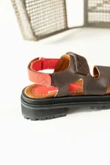 Sandalias de cuero marrón con diseño de tiras anchas y ajuste mediante velcro. Presentan una plantilla acolchada color tostado con detalles en rojo y una suela de goma negra con plataforma dentada.