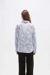 Camisa de algodón con diseño de rayas verticales finas en color celeste y blanco, con apliques florales bordados en relieve. Presenta cuello camisero, cierre frontal con botones y mangas largas con puños abotonados.