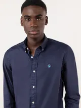 Camisa azul de hombre confeccionada en 100% algodón twill, tintada en prenda. Cuenta con corte regular fit, cuello button-down, cierre frontal de botones, manga larga con puño simple y pequeño logo rojo bordado en el pecho.