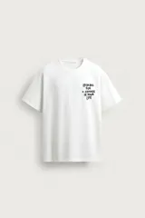 Camiseta blanca de corte relajado, con cuello redondo y manga corta. Presenta un pequeño estampado de texto en contraste en la parte frontal izquierda del pecho.