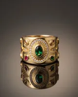 Anillo ancho de bronce con baño de oro, diseño texturizado con detalles ornamentales, piedra central verde ovalada rodeada de circonias y pequeñas piedras rojas incrustadas.