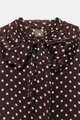 Blusa de manga larga, color marrón oscuro con estampado de lunares blancos. Presenta cuello alto con lazo ajustable en la espalda y puños elásticos.