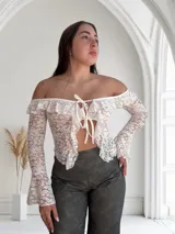 Top de encaje blanco con mangas largas acampanadas, hombros descubiertos, volados y lazo ajustable en el frente.