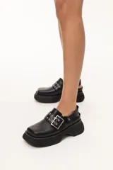 Zueco de cuero negro con plataforma gruesa, punta cuadrada y tira ancha con hebilla metálica y ojales decorativos.