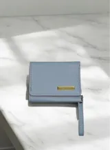 Billetera compacta de diseño plegable en color celeste, con textura granulada. Presenta una placa metálica dorada con el logo de la marca en el frente y una pequeña correa lateral del mismo tono.