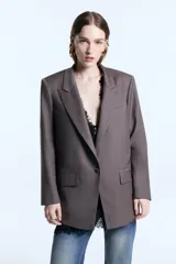 Blazer de corte oversize confeccionado en sarga, con hombreras marcadas, solapas clásicas y cierre frontal con un botón forrado a tono. Presenta dos bolsillos delanteros con solapa, logo bordado en una de las tapas y bordado decorativo en el cuello trasero. El diseño cuenta con un largo irregular que llega por debajo de la cadera y está completamente forrado.