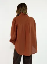 Camisa de algodón color blanco, con cuello redondo y detalle de volados en el cuello y puños. Presenta mangas largas con puños elásticos y un diseño de corte holgado con ruedo asimétrico, más largo en la parte trasera.