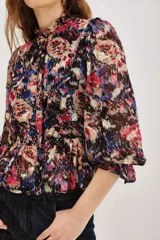 Camisa de manga larga con estampado floral y detalles en hilo metalizado. Presenta cuello alto con volados, cierre frontal con botones y puños con volados. El diseño incluye un corte entallado en la cintura con un ligero peplum.