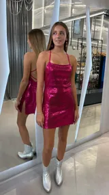 Vestido corto fucsia de lentejuelas, con escote drapeado, tirantes finos cruzados en la espalda y abertura lateral en la falda.