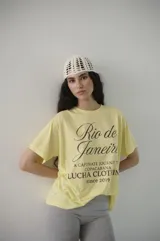 Remera de algodón color amarillo claro, con estampado frontal en marrón que dice "Rio de Janeiro A Captivate Journey Copacabana Lucha Clothin since 2019".