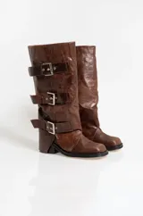 Botas de caña alta confeccionadas en cuero con acabado gastado. Presentan un diseño con tres correas horizontales ajustables mediante hebillas metálicas plateadas. Cuentan con un taco cuadrado bajo y suela de goma.