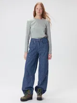 Pantalón de tiro medio y pierna ancha confeccionado en denim de algodón grueso. Presenta un diseño a rayas verticales en tonos azul y celeste, con costuras en contraste color rojo, bolsillos laterales tipo cargo y remaches metálicos.