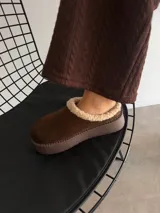 Sueco de estilo casual confeccionado en símil gamuza color marrón, con interior y borde superior revestido en corderito sintético color crema. Presenta una suela de plataforma plana de goma en tono marrón oscuro.