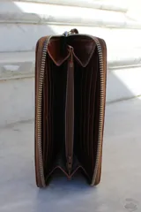 Billetera tipo sobre con correa de muñeca, confeccionada en cuero con textura de reptil en color marrón. Cuenta con cierre perimetral y placa metálica con el logo de la marca en el frente.