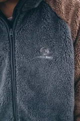 Campera con capucha de tejido sherpa, combinando paneles en color gris y marrón. Presenta cierre frontal completo, mangas largas y diseño holgado.