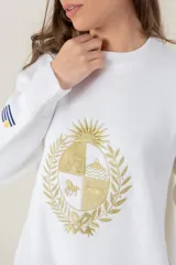 Buzo de tejido de punto color blanco, de corte holgado y cuello redondo. Presenta un bordado central en hilo dorado con el escudo nacional y un pequeño detalle bordado en la manga derecha con los colores de la bandera uruguaya.