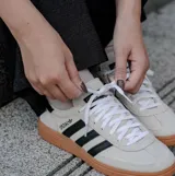 Championes Adidas modelo Handball Spezial, confeccionados en gamuza color beige con las icónicas tres tiras laterales en negro. Presentan suela de goma color caramelo y detalle de talón en negro.