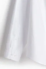 Camisa blanca de manga larga con cuello inglés, tapeta lisa y bajo redondeado. Presenta canesú en la espalda y puños ajustables con botón.