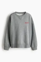 Buzo oversized de felpa color gris melange, con cuello redondo y hombros caídos. Cuenta con un pequeño bordado rojo con texto en el pecho y puños y bajo con ribete acanalado.