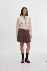 Camisa de tacto de seda color beige, entallada en la cintura, con cuello camisero, bolsillos frontales con solapa y bordados, y cordón decorativo en el cuello.