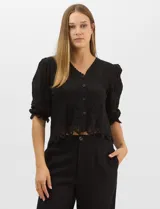 Blusa negra de corte recto con mangas al codo ligeramente abullonadas. Confeccionada en algodón con motivos florales bordados a tono, presenta escote en V y cierre frontal con botones. Incluye detalles de encaje calado estilo macramé en puños y ruedo con terminaciones en ondas.
