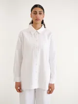 Camisa blanca de manga larga con cuello clásico y cierre frontal de botones, de corte recto y largo por debajo de la cadera.