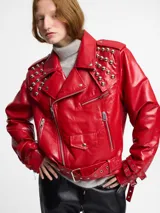 Chaqueta biker negra de cuero sintético con tachas plateadas en los hombros y solapas, cierre asimétrico con cremallera, cinturón ajustable con hebilla y detalles metálicos.