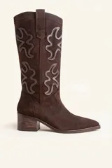 Bota de caña alta estilo tejano confeccionada en gamuza color marrón oscuro. Presenta detalles de costuras decorativas en forma de llama en la caña, taco cuadrado de madera y punta almendrada.