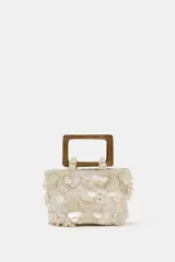 Bolso de mano formato mini confeccionado en tejido texturizado color crema. Presenta apliques de nácar y bordados florales en relieve, con doble asa rígida de madera y cadena bandolera extraíble.