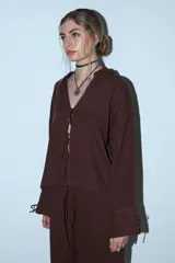 Camisa de manga larga color crema o beige claro, con textura arrugada, cuello camisero y cierre frontal mediante lazos anudados.