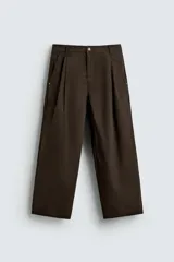 Pantalón chino de corte wide fit, color marrón oscuro, confeccionado en tejido de algodón. Presenta pliegues frontales, bolsillos laterales y bolsillos traseros de vivo, con cierre frontal de cremallera y botón.