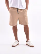 Bermuda de algodón en color beige claro, con cintura elástica ajustable mediante cordón y bolsillos laterales. Presenta un pequeño parche con logo en la parte inferior de la pierna.