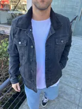 Chaqueta de jean color negro con cuello de corderito negro, cierre frontal con botones metálicos y bolsillos frontales con solapa.