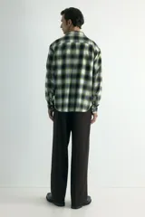 Camisa de franela de algodón suave con cuello inglés, cierre frontal de botones, mangas largas con puños abotonados y bajo redondeado. Presenta un diseño a cuadros en tonos oscuros y amarillentos.