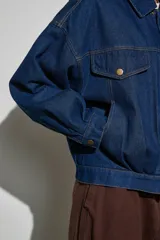 Campera de jean azul oscuro, de corte oversize, con cuello clásico, cierre frontal con cremallera y botones a presión, bolsillos frontales con solapa y mangas largas.