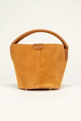 Mini cartera tipo balde o cubo, confeccionada en gamuza color camel o tabaco, con forro interior. Posee un asa superior de cuero liso marrón más oscuro y un cierre de solapa con broche en la parte superior central.