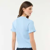 Camiseta de punto celeste con cuello redondo, mangas cortas y cierre frontal de botones, adornada con volantes en la botonadura, los puños y el dobladillo.